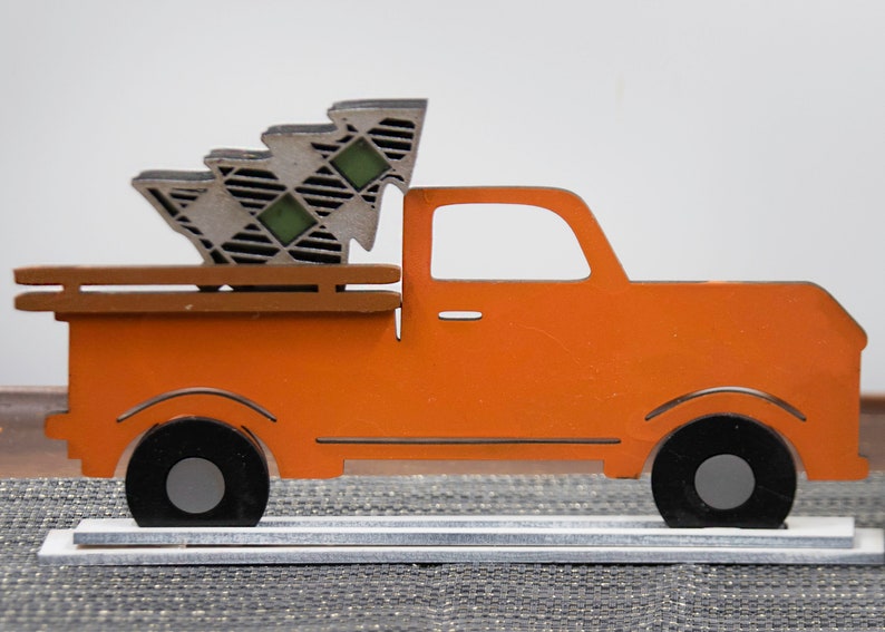 Puede incluir: Cami&oacute;n de madera naranja con un &aacute;rbol de Navidad a cuadros en la caja. El cami&oacute;n est&aacute; sobre una base de madera blanca.