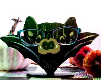 May include: A black bat-shaped glasses holder with a painted face and blue-tinted glasses. The holder is sitting on a black and white checkered surface with a white pumpkin and red pumpkins in the background.