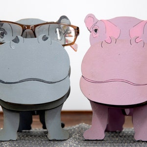 Grey and lavender wooden standing Hippos. Mouth / nose sticks out to hold glasses. Brown glasses on Grey hippos face. darker Grey mouth, nostrils, eyes and ears. lavender Hippo has pink mouth, nostrils, eyes & ears. Both standing on a desk & placemat