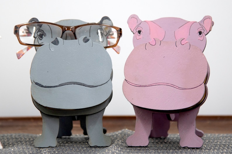 Grey and lavender wooden standing Hippos. Mouth / nose sticks out to hold glasses. Brown glasses on Grey hippos face. darker Grey mouth, nostrils, eyes and ears. lavender Hippo has pink mouth, nostrils, eyes & ears. Both standing on a desk & placemat