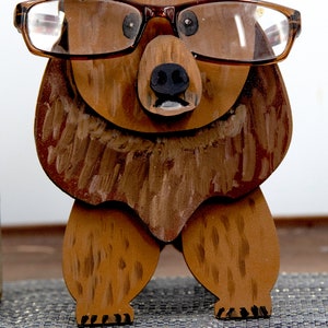 Wooden brown grizzley bear- brown glasses on its face. Brown body & head, darker steaks for fur, Darker brown chest & light streaks for texture. protruding nose holds glasses, black nose & mouth, black eyes white pupils. On desk on silver placemat