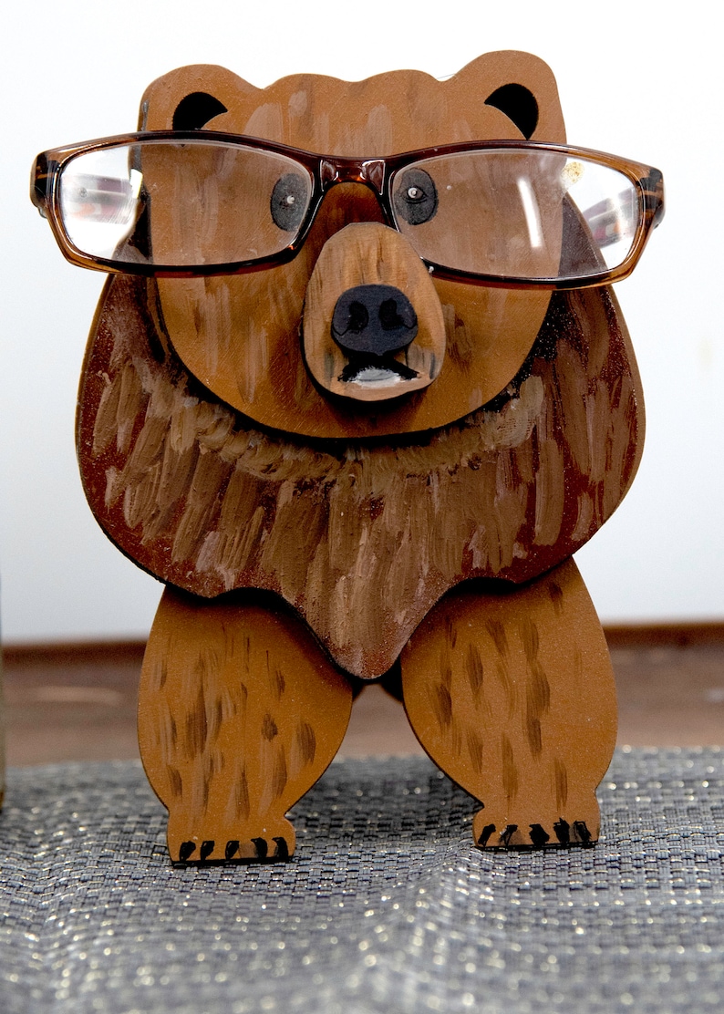 Wooden brown grizzley bear- brown glasses on its face. Brown body & head, darker steaks for fur, Darker brown chest & light streaks for texture. protruding nose holds glasses, black nose & mouth, black eyes white pupils. On desk on silver placemat
