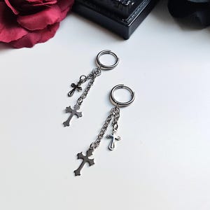 Könnte beinhalten: Ein Paar silberfarbene Creolen mit Kreuzanhängern. Jede Ohrring hat einen kleinen Reifen, eine Kette und drei Kreuzanhänger in verschiedenen Größen. Die Ohrringe liegen auf einer weißen Oberfläche, im Hintergrund eine dunkelrote Rose.