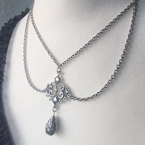 May include: A silver head chain necklace with an ornate centerpiece and a teardrop pendant. The centerpiece has a symmetrical design with swirling details. The necklace features two chain strands that drape across the forehead.