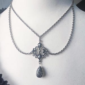 May include: Silver necklace featuring a two-strand chain design. The central pendant is an ornate, symmetrical piece with a teardrop charm. The necklace has a vintage aesthetic, suitable for gothic or bohemian styles.