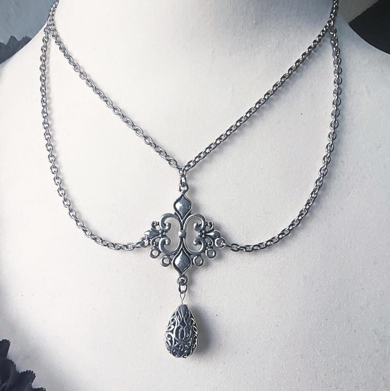 May include: Silver necklace featuring a two-strand chain design. The central pendant is an ornate, symmetrical piece with a teardrop charm. The necklace has a vintage aesthetic, suitable for gothic or bohemian styles.