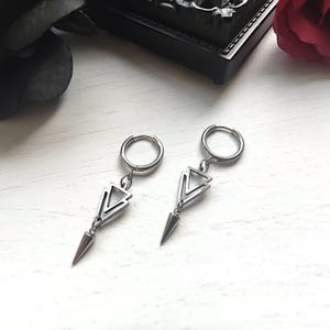 May include: A pair of silver-colored hoop earrings. Each earring features a geometric design with a triangle and a pointed, cone-shaped dangle. The earrings are displayed on a white, textured surface.