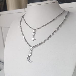 Può includere: Una collana d'argento a due fili con un ciondolo a forma di stella e mezzaluna. La collana è esposta su un manichino bianco. La catena è un delicato design a maglie e i ciondoli sono semplici ed eleganti.