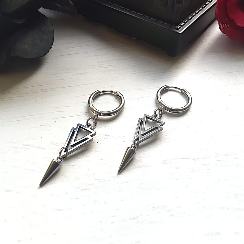 May include: A pair of silver-toned earrings. Each earring features a small hoop that connects to a geometric design with three stacked triangles and a pointed, cone-shaped dangle. The earrings are displayed on a white wooden surface.