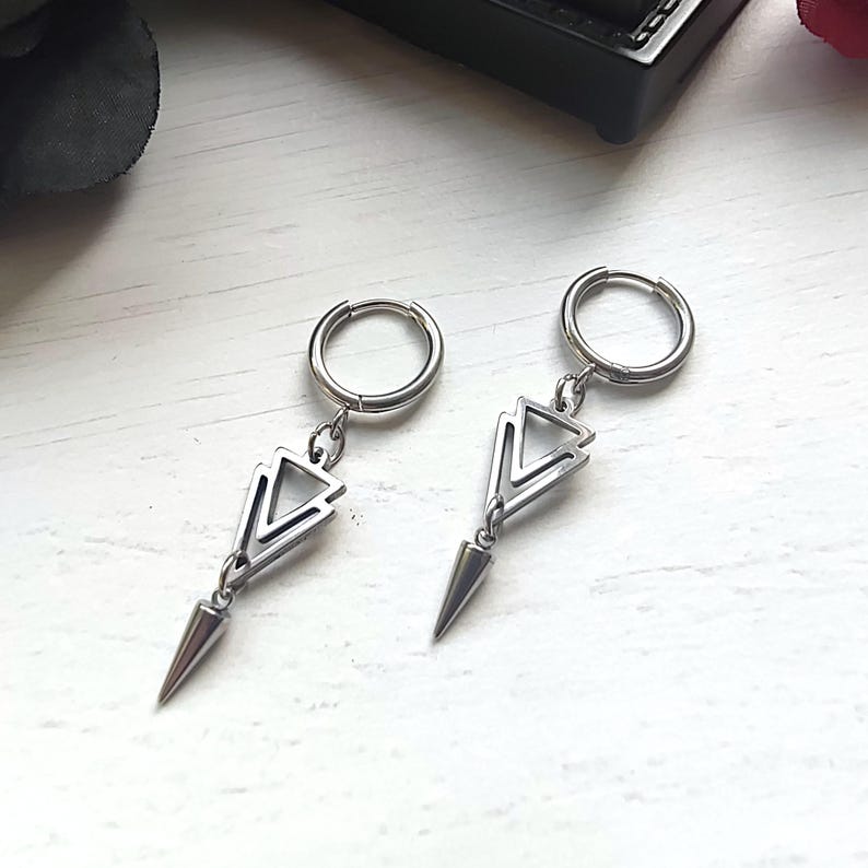 May include: A pair of silver-colored earrings. Each earring features a small hoop that connects to a geometric triangle design, which is connected to a cone-shaped spike. The earrings are displayed on a white surface.