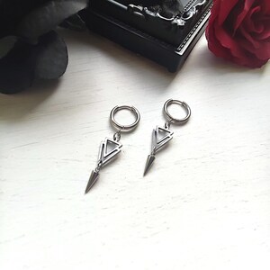 May include: A pair of silver-colored hoop earrings. Each earring features a geometric design with a triangular shape and a pointed, cone-shaped dangle. The earrings are displayed on a white surface, with a dark background.