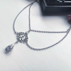 May include: Silver headpiece with a delicate chain design. The headpiece features an ornate centerpiece with a teardrop-shaped pendant. The chains are connected to the centerpiece and are designed to be worn across the forehead and around the head.