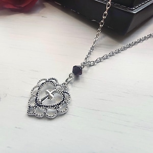 May include: Silver necklace with a heart-shaped pendant and a cross. The pendant has a filigree design and a small, dark purple bead. The chain is delicate. The necklace is displayed on a light-coloured surface.