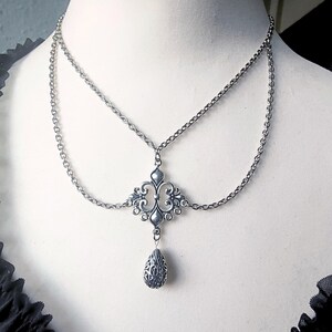 May include: Silver-coloured, two-strand necklace with an ornate, symmetrical pendant. The pendant features a central design with swirling details and a teardrop-shaped charm. The necklace is displayed on a mannequin.