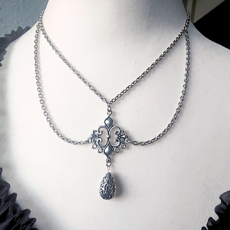 May include: Silver-coloured, two-strand necklace with an ornate, symmetrical pendant. The pendant features a central design with swirling details and a teardrop-shaped charm. The necklace is displayed on a mannequin.