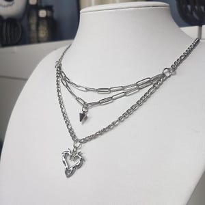 以下が含まれることがあります： シルバーカラーのレイヤードネックレス。2種類の異なるチェーンで構成されています。上段は長方形のリンクチェーンに小さな円錐形のペンダントが付き、下段は標準的なチェーンにハート型のチャームが付いています。