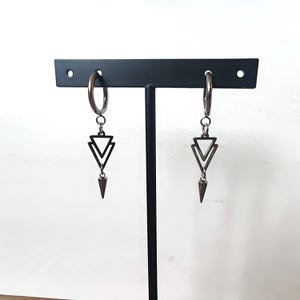 May include: A pair of silver hoop earrings with a geometric design. Each earring features a double triangle pendant and a pointed, cone-shaped charm. The earrings are displayed on a black stand against a white background.