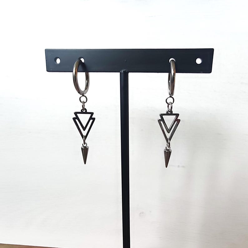 May include: A pair of silver hoop earrings with a geometric design. Each earring features a double triangle pendant and a pointed, cone-shaped charm. The earrings are displayed on a black stand against a white background.