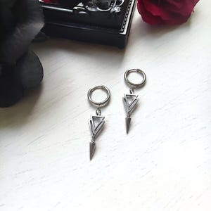 May include: A pair of silver-colored earrings. Each earring features a circular hoop, a triangular design, and a pointed, cone-shaped dangle. The earrings are displayed on a white surface, with a dark background and a red rose in the background.