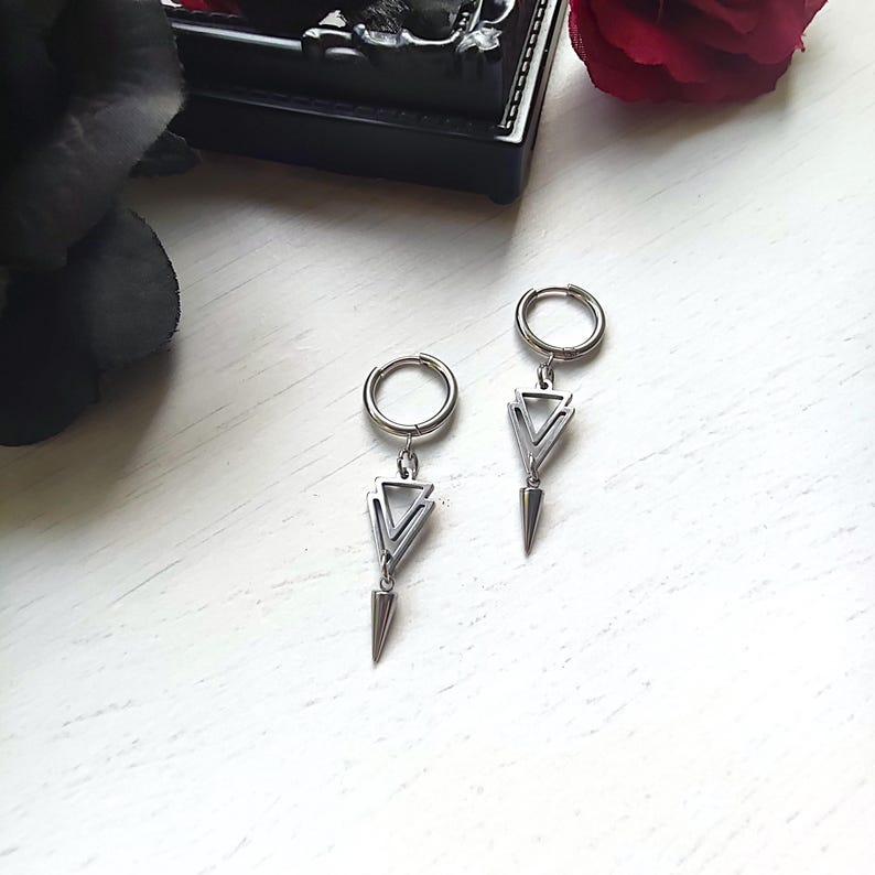 May include: A pair of silver-colored earrings. Each earring features a circular hoop, a triangular design, and a pointed, cone-shaped dangle. The earrings are displayed on a white surface, with a dark background and a red rose in the background.