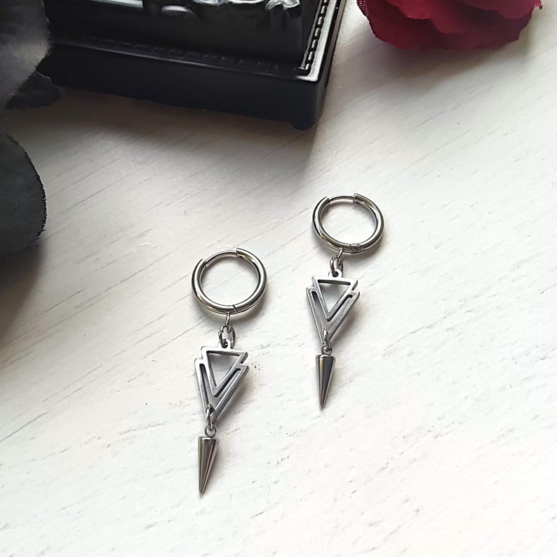 May include: A pair of silver-colored hoop earrings. Each earring features a geometric triangle design with a pointed bottom. The earrings are displayed on a white surface, with a dark object and a red rose in the background.