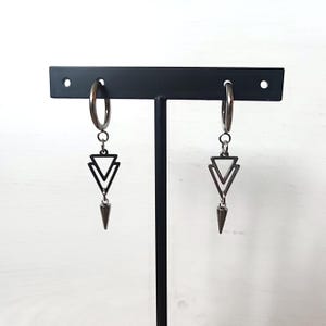 May include: A pair of silver hoop earrings with a geometric design. Each earring features a double triangle pendant and a pointed cone charm. The earrings are displayed on a black stand against a white background.