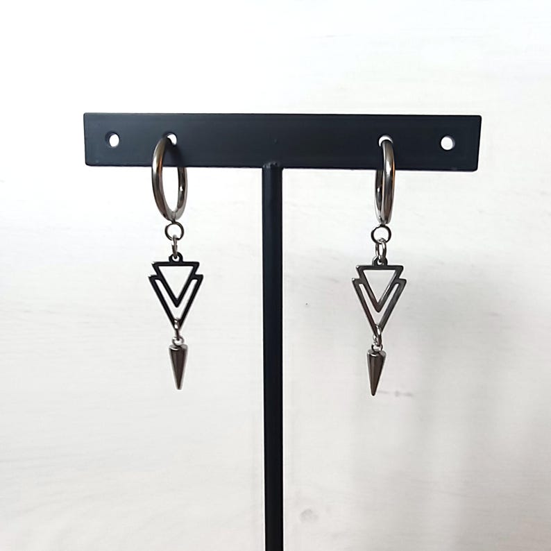 May include: A pair of silver hoop earrings with a geometric design. Each earring features a double triangle pendant and a pointed cone charm. The earrings are displayed on a black stand against a white background.