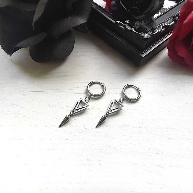 May include: A pair of silver-toned earrings. Each earring features a small hoop at the top, connected to a geometric triangle design, and a pointed, elongated teardrop shape. The earrings are displayed on a white surface, with dark floral accents in the background.