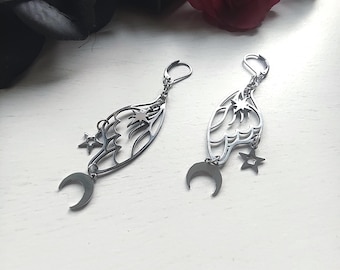 May include: Silver-toned dangle earrings featuring a celestial design. Each earring has a wing-shaped filigree, a crescent moon, and star charms. The earrings are displayed on a white surface with dark floral accents.