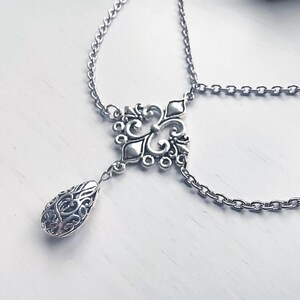 May include: Silver-coloured body jewellery featuring an ornate, filigree-style pendant with a teardrop-shaped charm. The jewellery has a delicate chain design and is displayed on a white surface. The piece is likely designed for the wrist or ankle.