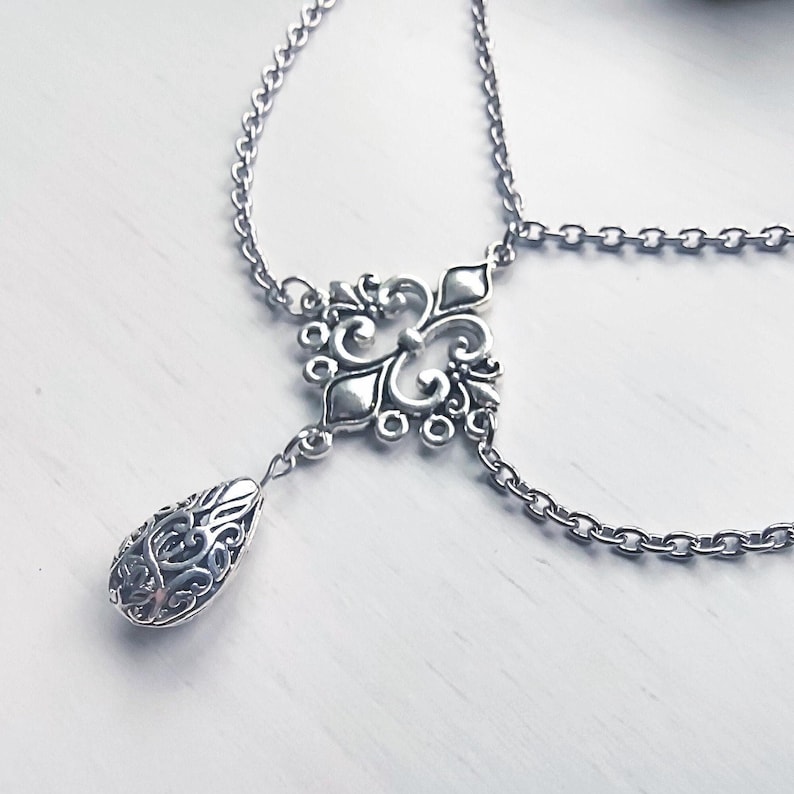May include: Silver-coloured body jewellery featuring an ornate, filigree-style pendant with a teardrop-shaped charm. The jewellery has a delicate chain design and is displayed on a white surface. The piece is likely designed for the wrist or ankle.