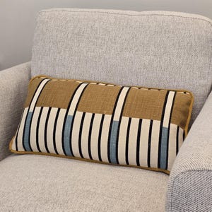 Puede incluir: Cojín decorativo rectangular con un patrón geométrico. El cojín presenta un fondo beige con rayas verticales negras, blancas y azul claro. Tiene un ribete dorado y está colocado sobre un sillón gris claro.