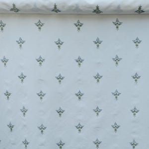May include: White fabric with a repeating pattern of stylized floral bouquets in shades of blue and green. The fabric is rolled up at the top, showing the pattern's continuity. The design is evenly spaced across the material.