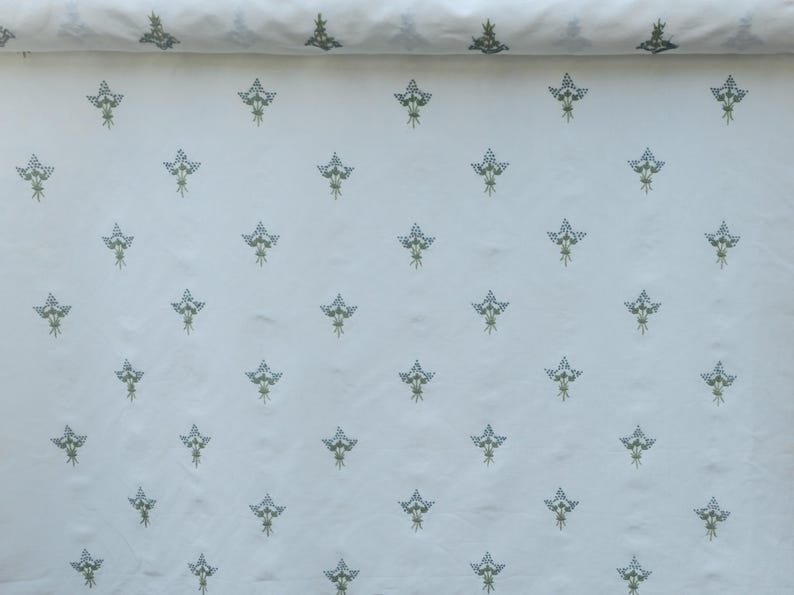 May include: White fabric with a repeating pattern of stylized floral bouquets in shades of blue and green. The fabric is rolled up at the top, showing the pattern's continuity. The design is evenly spaced across the material.