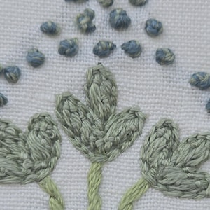 May include: Close-up of white fabric with embroidered floral design. The embroidery features three stylized green leaves with green stems and small blue and gray textured accents, creating a delicate, handcrafted look.