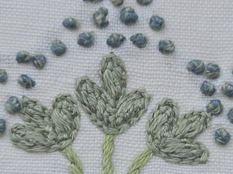May include: Close-up of white fabric with embroidered floral design. The embroidery features three stylized green leaves with green stems and small blue and gray textured accents, creating a delicate, handcrafted look.