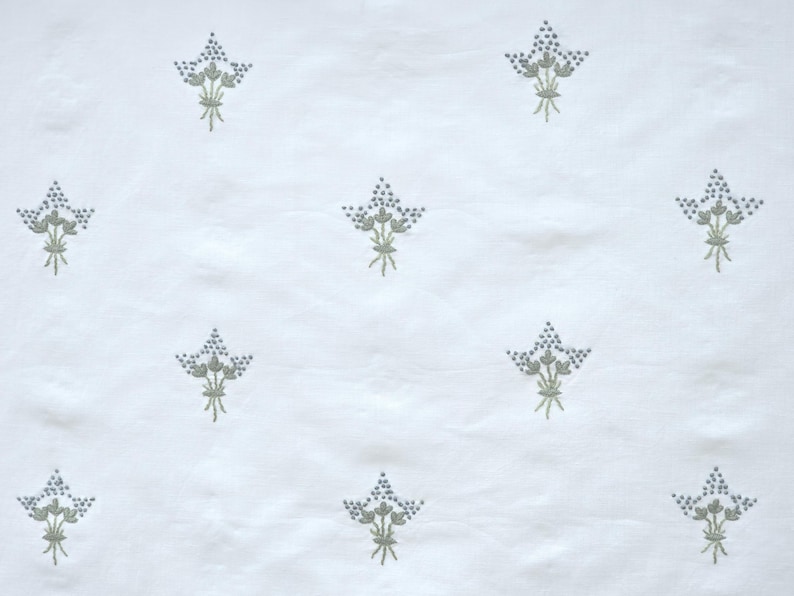 May include: White fabric with a repeating pattern of embroidered floral bouquets. Each bouquet features small, blue-gray flower clusters and green stems, creating a delicate and textured design.