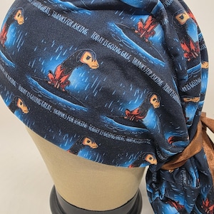 May include: A navy blue scrub cap featuring a repeating pattern of a creature with orange and black details, set against a blue and white background. The cap has the text "TODAY IS GOING GREAT, THANKS FOR ASKING" in white. A brown tie is attached.