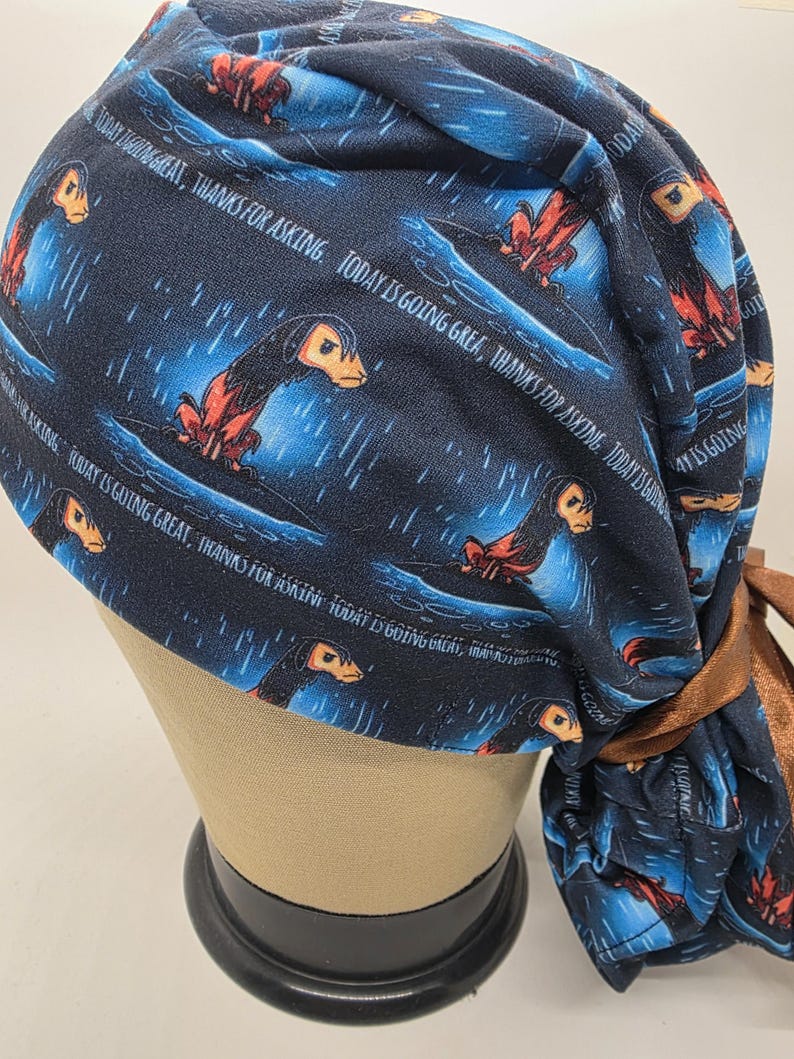 May include: A navy blue scrub cap featuring a repeating pattern of a creature with orange and black details, set against a blue and white background. The cap has the text "TODAY IS GOING GREAT, THANKS FOR ASKING" in white. A brown tie is attached.