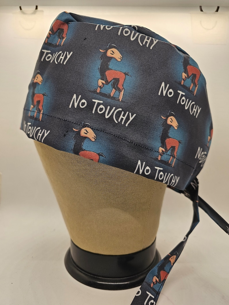 May include: A dark blue scrub cap with a repeating pattern of a cartoon llama and the words "No Touchy" in white. The llama is brown and red. The cap is tied at the back with matching fabric ties.