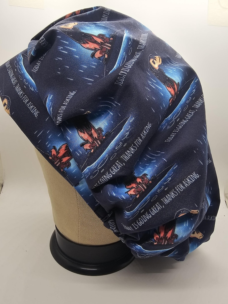 May include: Navy blue scrub cap with a repeating pattern of a cartoon character in a boat, with the text "TODAY IS GOING GREAT, THANKS FOR ASKING." The cap is made of fabric and is designed to cover the head.