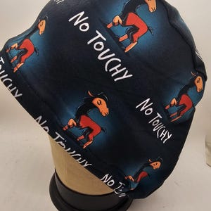 May include: A dark blue scrub cap featuring a repeating pattern of a cartoon horse with orange and black coloring. The words "No Touchy" are written in white. The cap is displayed on a mannequin head.