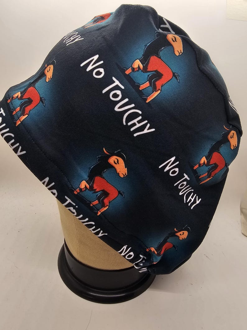 May include: A dark blue scrub cap featuring a repeating pattern of a cartoon horse with orange and black coloring. The words "No Touchy" are written in white. The cap is displayed on a mannequin head.