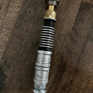 May include: A cylindrical prop constructed from industrial metal components, featuring a silver-toned threaded base, a central section with black ribbed rings, a brass-colored connector, and a silver-toned top cap with a circular grooved design.