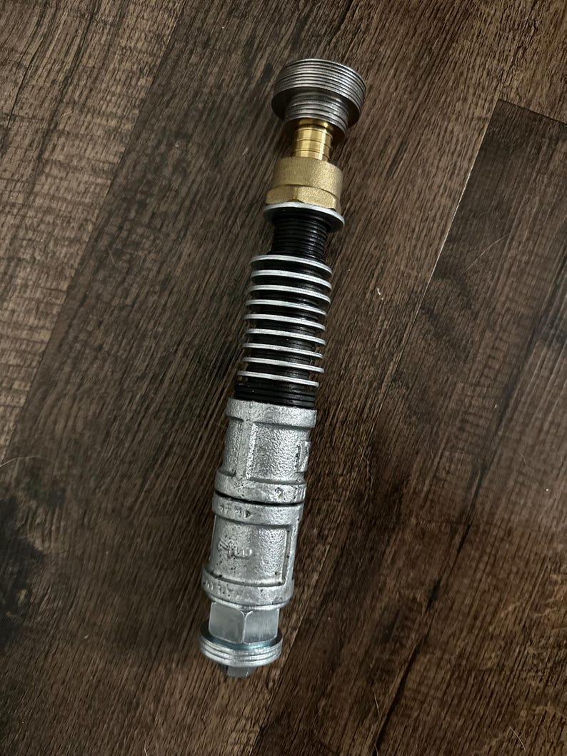 May include: A cylindrical prop constructed from industrial metal components, featuring a silver-toned threaded base, a central section with black ribbed rings, a brass-colored connector, and a silver-toned top cap with a circular grooved design.
