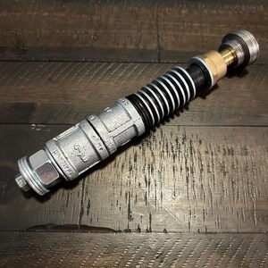 May include: A cylindrical prop constructed from industrial metal plumbing components, featuring a textured silver-colored pipe section, a series of black and silver ribbed rings, a brass-toned connector, and a threaded silver end cap.