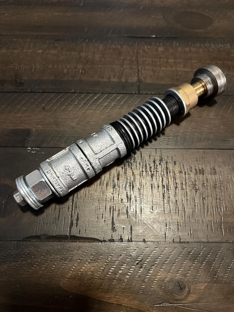 May include: A cylindrical prop constructed from industrial metal plumbing components, featuring a textured silver-colored pipe section, a series of black and silver ribbed rings, a brass-toned connector, and a threaded silver end cap.