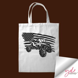 May include: A white tote bag with a black silhouette of an off-road vehicle driving over a distressed American flag.