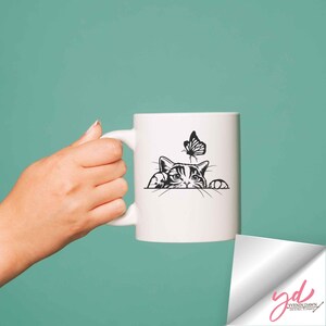 Pode incluir: Caneca de cer&acirc;mica branca com uma ilustra&ccedil;&atilde;o em preto e branco de um gato espiando pela borda da caneca e uma borboleta voando sobre o gato.