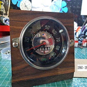 Könnte beinhalten: Eine Holzuhr mit einem Vintage-Auto-Tachometer-Zifferblatt. Das Zifferblatt der Uhr ist schwarz mit weißen Zahlen und einem roten Zeiger. Die Uhr ist auf einem dunklen Holzsockel montiert. Der Text "1960 - 1967 BEETLE" ist auf einem beigefarbenen Etikett unter der Uhr gedruckt.